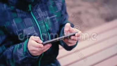 男孩坐在公园里的长凳上用手机玩游戏。 特写男孩`手。